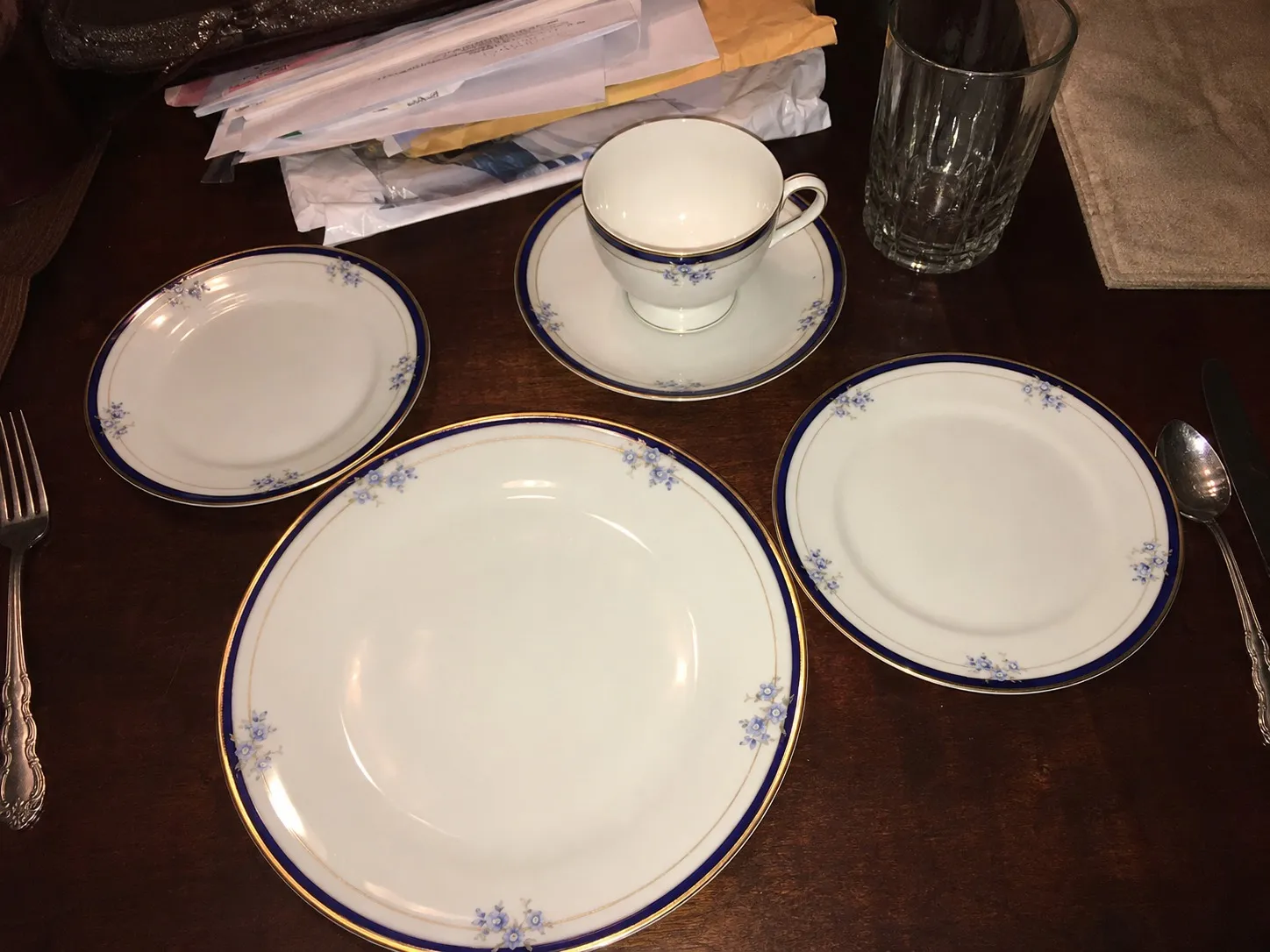 A five-piece china place setting photographed on a dining table under a ceiling light, the mail pushed to one side