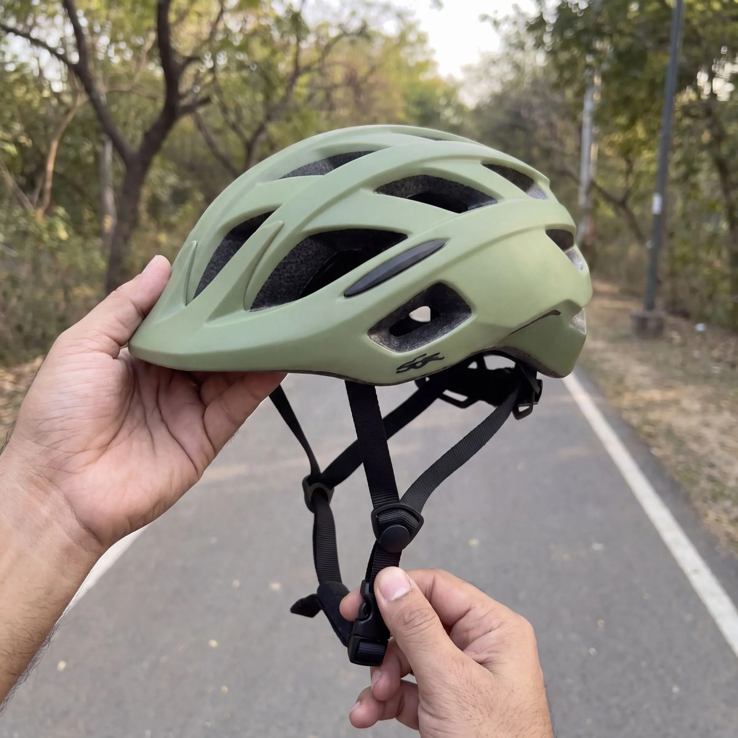 An olive bicycle helmet, held up by its strap, a road behind it
